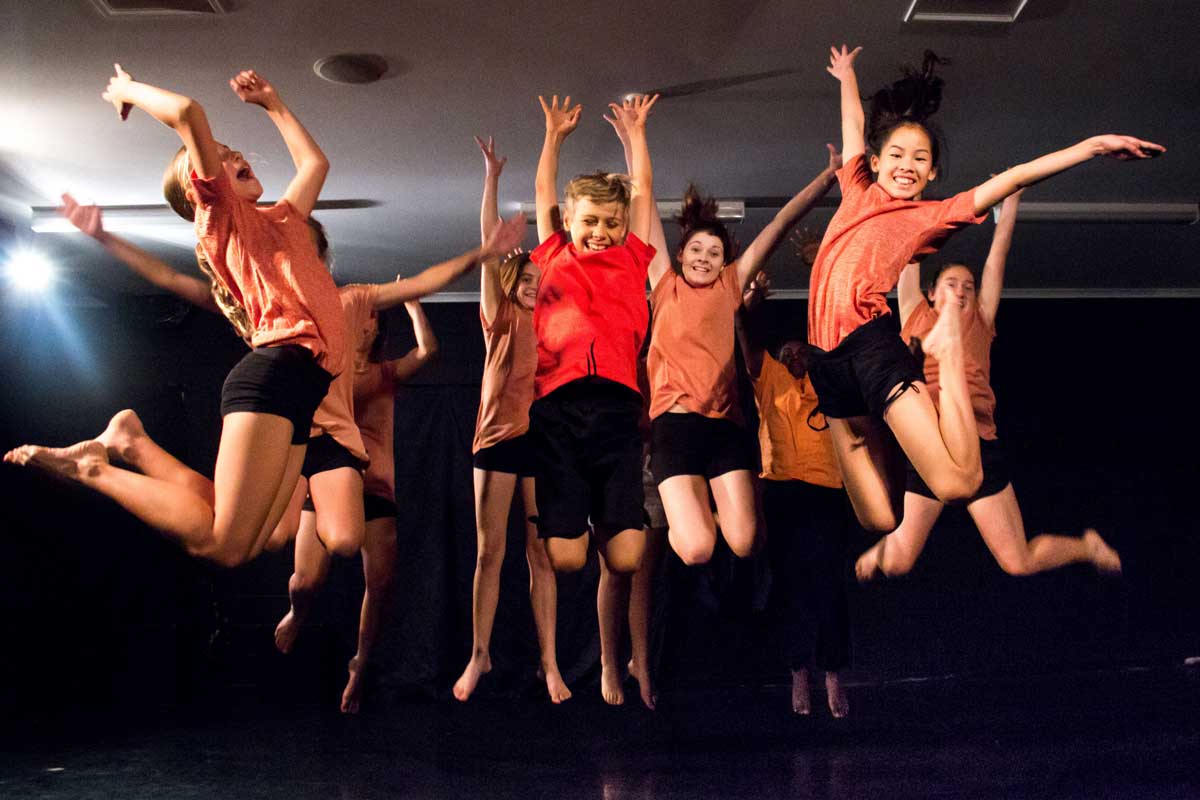 A group of young people all jumping up with their arms outstretched above their heads, smiles on their faces. They are in a room with lights on.